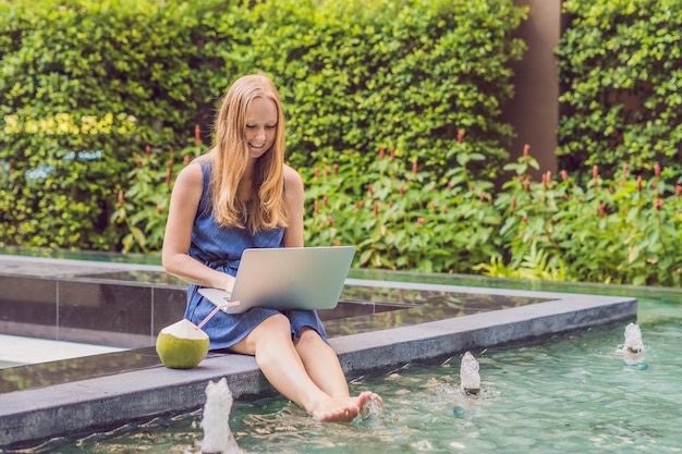 Freelancer mujer joven sentada cerca de la piscina con su computadora portátil en el hotel navegando en su teléfono inteligente. Ocupado en vacaciones. Concepto de trabajo distante. Copie el espacio para su texto