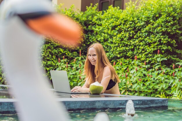 Freelancer mujer joven sentada cerca de la piscina con su computadora portátil en el hotel navegando en su teléfono inteligente. Ocupado en vacaciones. Concepto de trabajo distante. Copie el espacio para su texto