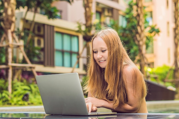 Freelancer mujer joven sentada cerca de la piscina con su computadora portátil en el hotel navegando en su teléfono inteligente. Ocupado en vacaciones. Concepto de trabajo distante. Copie el espacio para su texto