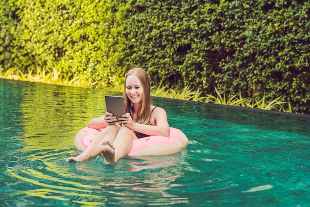 Freelancer mujer joven sentada cerca de la piscina con su computadora portátil en el hotel navegando en su teléfono inteligente. Ocupado en vacaciones. Concepto de trabajo distante. Copie el espacio para su texto.