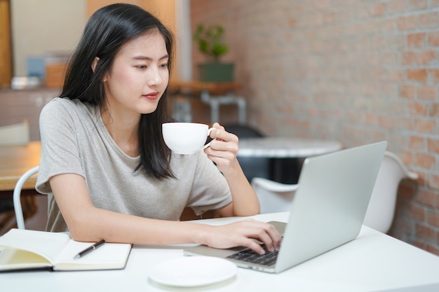 Freelancer novo asiático que usa o portátil e trabalhando quando escritório do café da bebida em casa.