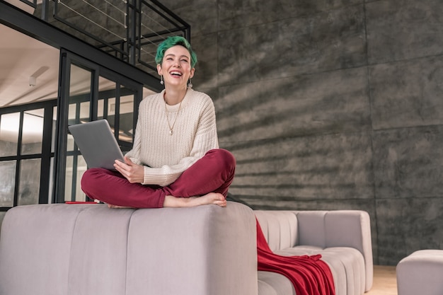 Freelancer rindo. Freelancer de cabelos verdes sentado no sofá rindo enquanto trabalha em um novo projeto