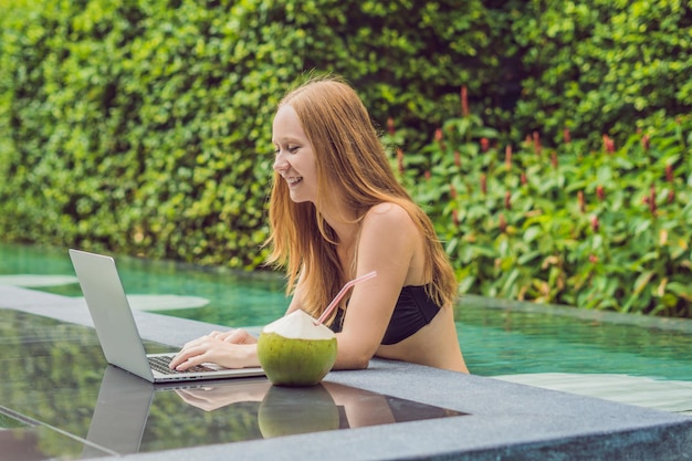 Freelancer de sexo femenino joven que se sienta cerca de la piscina con su computadora portátil. Ocupado en vacaciones. Concepto de trabajo distante. Copie el espacio para su texto.