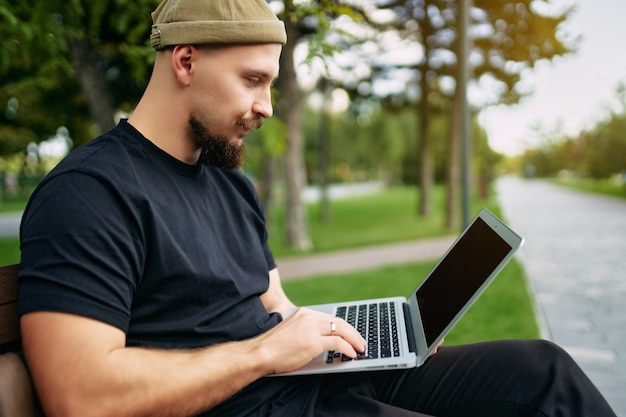 Freelancer se sienta en el parque de la ciudad mientras escribe en la computadora portátil blogger hipster viajero joven elegante