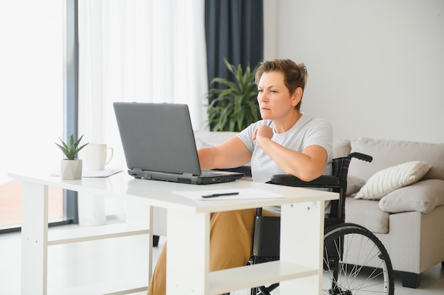 Freelancer en silla de ruedas usando una computadora portátil cerca de un cuaderno y papeles en la mesa