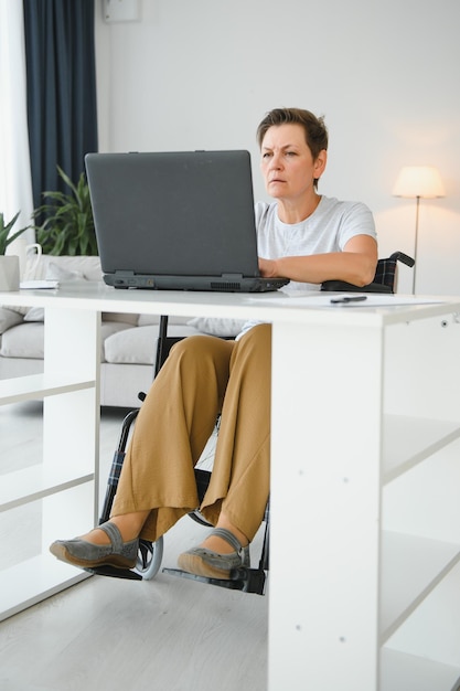 Freelancer en silla de ruedas usando una computadora portátil cerca de un cuaderno y papeles en la mesa