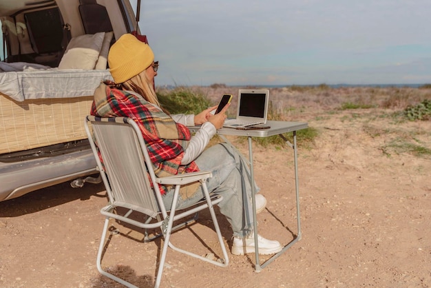 Freelancer trabalhando em um laptop sentado do lado de fora de um trailer falando pelo celular em um local costeiro tranquilo