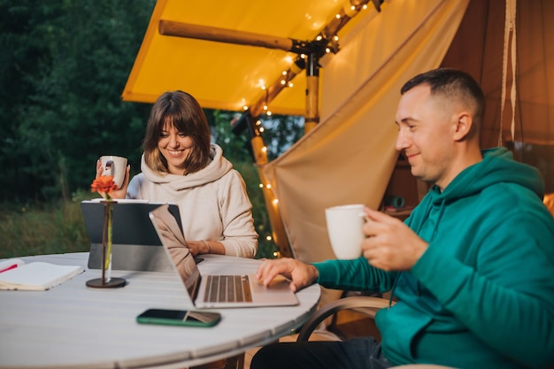 Freelancers de casal de família feliz bebem café enquanto trabalham laptop na aconchegante barraca de glamping na noite de verão Barraca de acampamento de luxo para férias ao ar livre e férias Conceito de estilo de vida