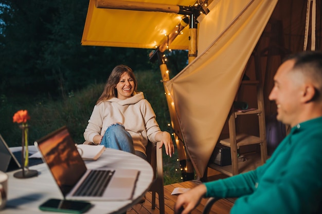 Freelancers de casal de família feliz descansam depois de trabalhar laptop na aconchegante barraca de glamping na noite de verão Barraca de acampamento de luxo para férias ao ar livre e férias Conceito de estilo de vida