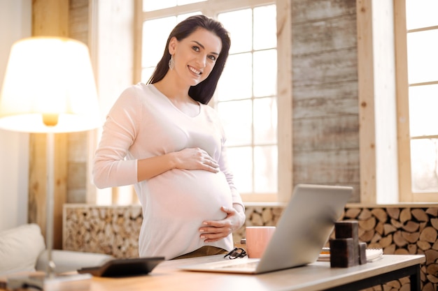 Freelancing durante a gravidez. Feliz grávida sincera abraçando a barriga enquanto olha para a câmera e se prepara para trabalhar