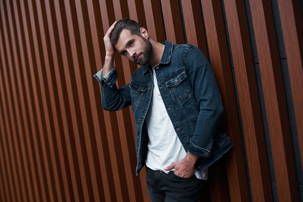 Foto freestyle joven de pie apoyado en una pared de madera aislado tocando el cabello mirando a un lado pensativo