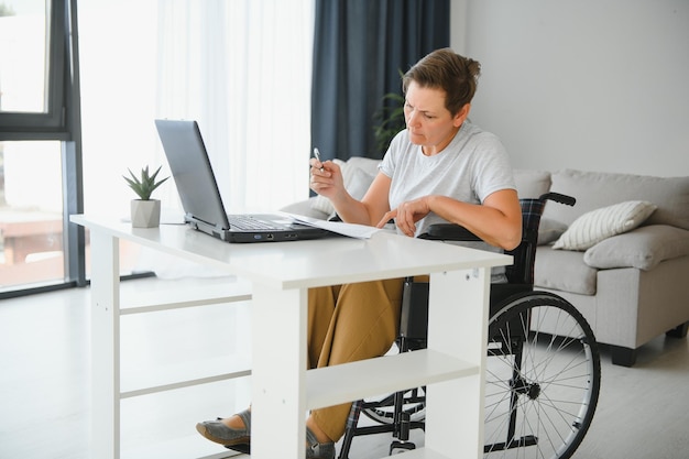 Freiberufler im Rollstuhl mit Laptop in der Nähe von Notizbuch und Papieren auf dem Tisch