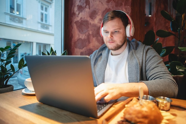 Freiberufler mit Headset, der im Café am Laptop arbeitet, Burger isst und Tee trinkt, Kopienraum