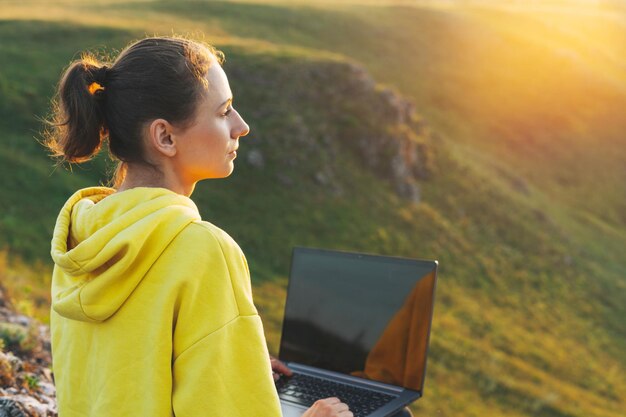 Freiberuflerin der jungen Frau im gelben Hoodie mit geöffnetem Laptop auf dem Hintergrund der schönen Aussicht