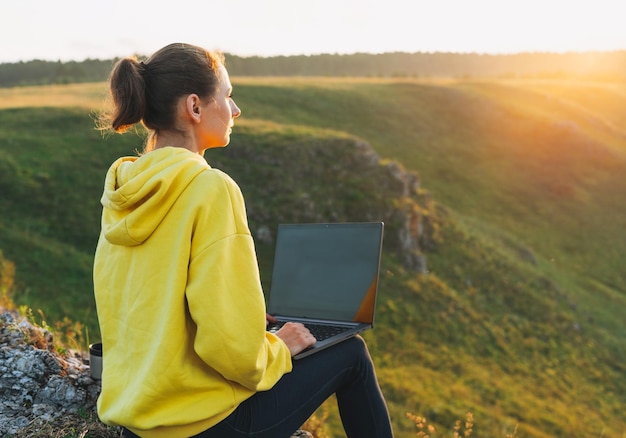 Freiberuflerin der jungen Frau im gelben Hoodie mit geöffnetem Laptop auf dem Hintergrund der schönen Aussicht