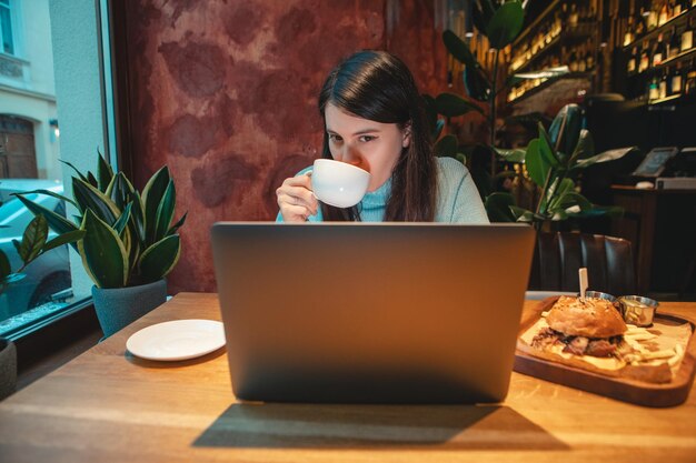 Freiberuflerin, die am Laptop im Café arbeitet und Tee trinkt, der Burger-Kopienraum isst