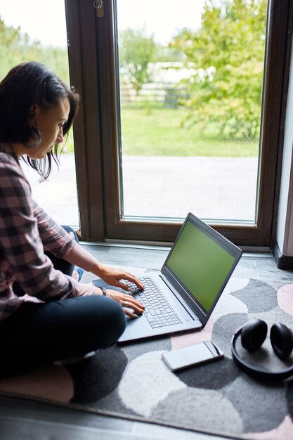 Freiberuflerin, die auf dem Laptop tippt, der zu Hause in der Nähe großer Fenster auf dem Boden sitzt, zufriedene Frau, die im Notebook surft, studiert, von zu Hause aus arbeitet.