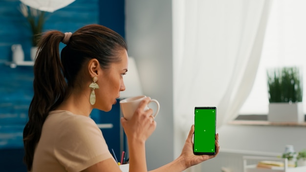 Freiberuflerin, die Kaffee im Schreibtisch trinkt und mit Kollegen spricht, indem sie ein Greenscreen-Chroma-Key-Smartphone verwendet. Kaukasische Frauen, die Online-Informationen suchen, verwenden isoliertes Telefon