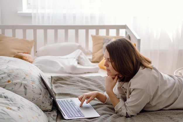Freiberuflerin für berufstätige Mutter Eine junge Mutter arbeitet am Laptop, während ihr Kleinkind auf der Krippe schläft