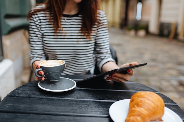 Freiberuflerin mit braunen Haaren, die an einem digitalen Tablet arbeitet, während sie sich auf der Café-Terrasse ausruht