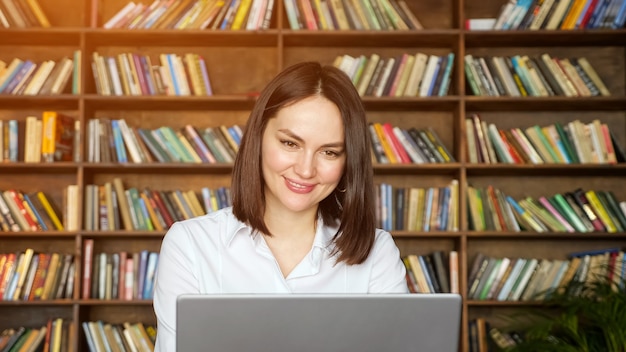 Freiberuflerin mit kurzen Haaren spricht online auf einem grauen Laptop gegen große Bücherregale in der örtlichen Bibliothek, Sonnenlicht