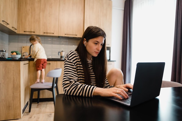 Freiberuflerin sitzt am Tisch im Büro der Küche zu Hause und arbeitet am Laptop Verspieltes Kind lenkt vom Arbeitskind ab, das Lärm macht und die Aufmerksamkeit der beschäftigten Mutter erbittet