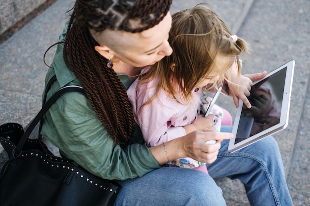 Freiberufliche Eltern. Vielbeschäftigte berufstätige Mutter mit kleiner Tochter geht auf der Straße spazieren und schaut auf ein Tablet. Multitasking. Freiberufliche Mutter macht sich Notizen, während sie ihr Baby draußen hält