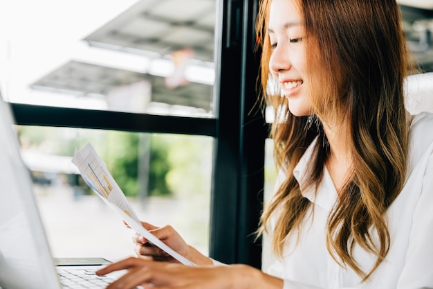 Freiberufliche Frau hält Papierkram dokumentiert Papierdiagramme und verwendet Laptop im Café, asiatische junge Geschäftsfrau, die Finanzen für Budget analysiert oder Diagrammpapier mit Computer berichtet