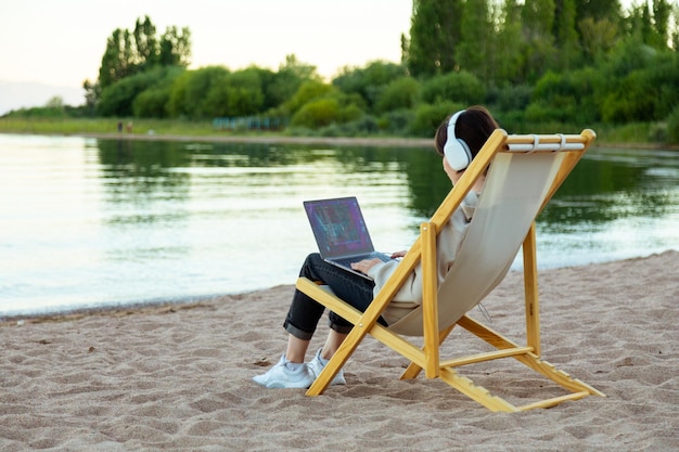 Freiberufliche Frau mit Kopfhörer sitzt seitwärts und arbeitet am Laptop am See
