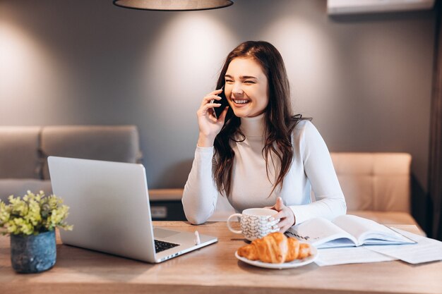 Freiberufliche Frau mit Laptop beim Sitzen zu Hause. Junge Frau, die in der Küche sitzt und am Laptop arbeitet. Hübsche Frau, die am Telefon spricht und während der Arbeit Tee-Kaffee trinkt.