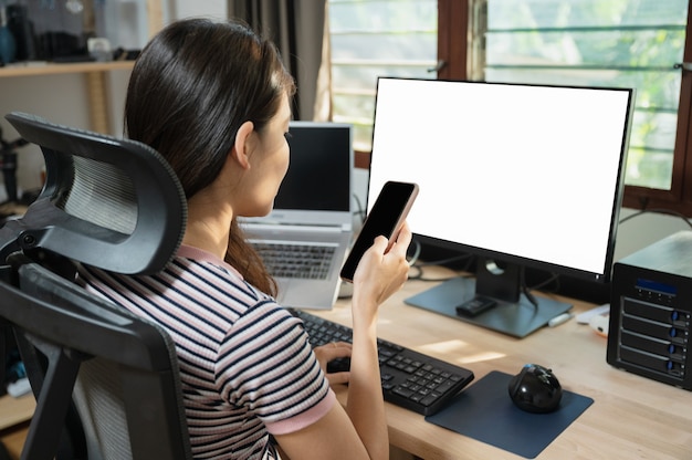 Freiberufliche junge Asiatin mit Handy am Arbeitstisch mit leerem Monitor im Haus