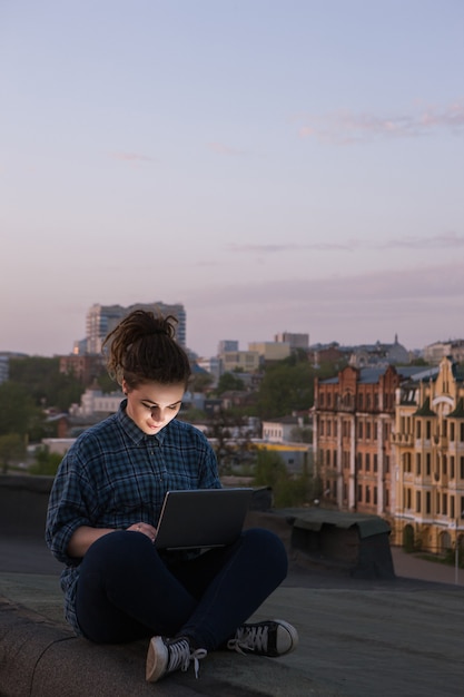 Freiberufliche Tätigkeit im Freien. Selbstbildung für Mädchen. Glückliche junge Frau mit Laptop auf dem Dach im Vordergrund, moderne Social-Media-Kommunikation, städtischer Hintergrund