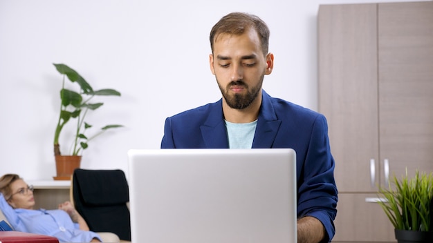 Freiberuflicher Geschäftsmann, der am Laptop im Haus arbeitet, und seine Frau ist im Hintergrund