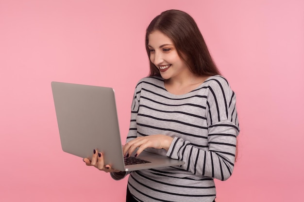 Freiberuflicher Job Internetkommunikation Porträt einer glücklichen jungen Frau in gestreiftem Sweatshirt, die auf einem Tastatur-Laptop tippt und lächelnd online chattet, Indoor-Studioaufnahme isoliert auf rosa Hintergrund