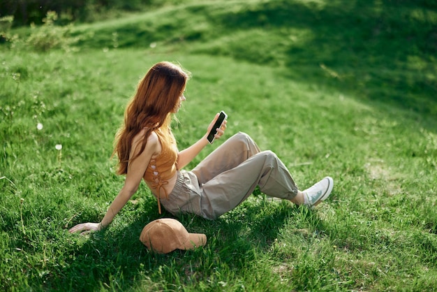 Freiberuflicher Student, der am Telefon arbeitet und im Sommer draußen auf grünem Gras sitzt, sein Handy und das Internet außerhalb der Stadt nutzt