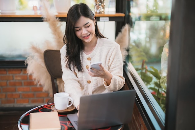 Freiberufliches Geschäft schönes positives Lächeln junge asiatische Frau, die warme Kleidung trägt, die online mit Nachricht auf dem Smartphone mit Laptop-Computer und Kaffeetasse im Haus im Wohnzimmer oder Café arbeitet