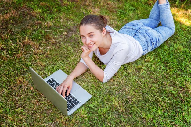 Freiberufliches Geschäftskonzept. Junge Frau, die auf grünem Rasen im Stadtpark liegt und an Laptop-PC-Computer arbeitet. Lifestyle authentische offene Studentin, die im Freien studiert. Mobiles Büro