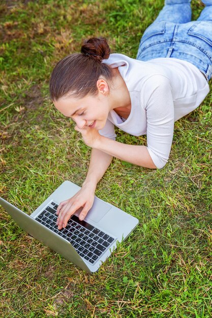 Freiberufliches Geschäftskonzept. Junge Frau, die auf grünem Rasen im Stadtpark liegt und an Laptop-PC-Computer arbeitet. Lifestyle authentische offene Studentin, die im Freien studiert. Mobiles Büro