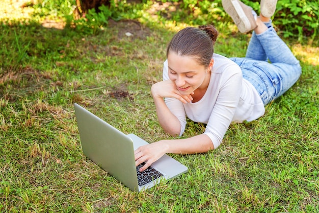 Freiberufliches Geschäftskonzept. Junge Frau, die auf grünem Rasen im Stadtpark liegt und an Laptop-PC-Computer arbeitet. Lifestyle authentische offene Studentin, die im Freien studiert. Mobiles Büro