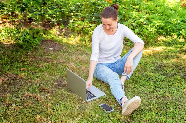 Freiberufliches Geschäftskonzept. Junge Frau sitzt auf grünem Rasen im Stadtpark und arbeitet an Laptop-PC-Computer. Lifestyle authentische offene Studentin, die im Freien studiert. Mobiles Büro