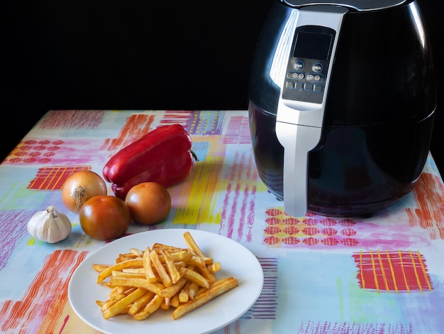 Freidora de aire cerrada sobre una mesa con papas fritas tomates pimientos cebolla y ajo Concepto de comida saludable