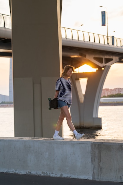 Freiheit Lifestyle Frau mittleren Alters Spaziergang in der Nähe des Flusses halten Longboard über Großstadtansicht bei Sonnenuntergang