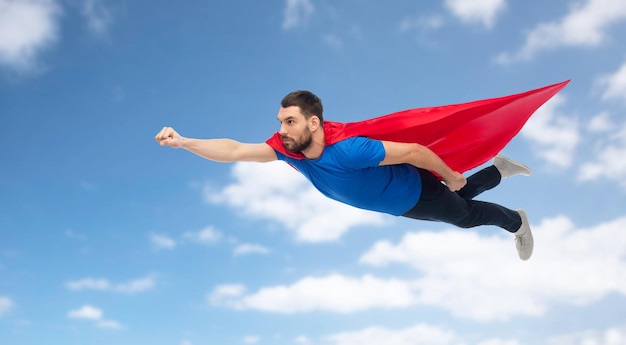 Freiheits-, Macht-, Bewegungs- und Menschenkonzept - Mann im roten Superheldenumhang, der über blauen Himmel und Wolkenhintergrund fliegt