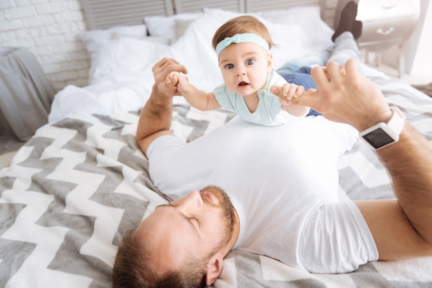 Freizeit. Beteiligt lächelndes hübsches Kleinkindmädchen, das auf ihrem Vater im Schlafzimmer liegt und beim Entspannen lächelt