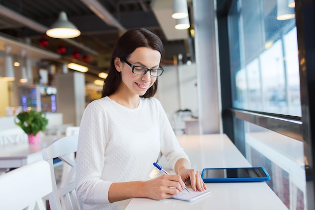 Freizeit-, Business-, People-, Technologie- und Lifestyle-Konzept - lächelnde junge Frau mit Brille mit Tablet-PC-Computer und Notebook, die sich im Café Notizen macht