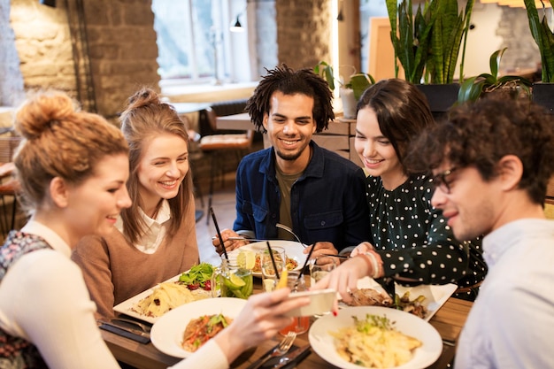 Freizeit-, Essens-, Getränke-, Menschen- und Urlaubskonzept - glückliche Freunde mit Smartphone beim Essen und Trinken im Restaurant