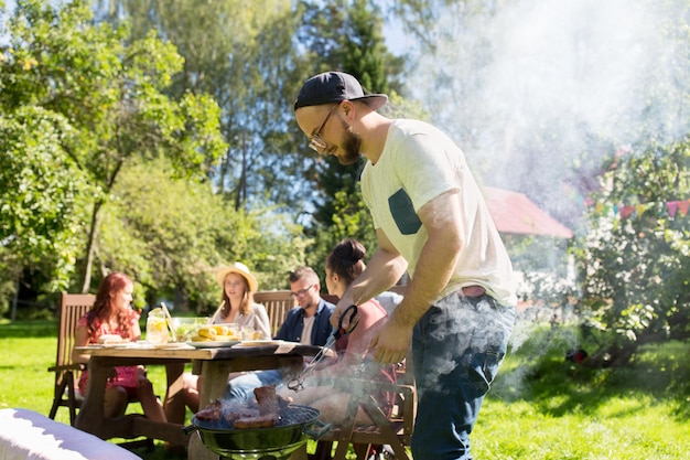 Freizeit-, Essens-, Menschen- und Urlaubskonzept - Mann kocht Fleisch auf dem Grill für seine Freunde auf einer Sommerparty im Freien
