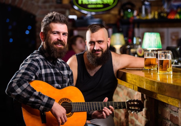 Freizeit für echte Männer. Hipster mit brutalem Bart verbringt Freizeit mit Freund in Bar. Mann spielt Gitarre in der Bar. Fröhliche Freunde entspannen sich bei Gitarrenmusik. Freitag Entspannung in der Bar. Freunde, die sich in der Bar oder im Pub entspannen.