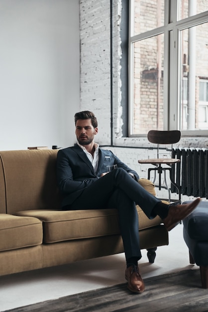 Foto freizeit genießen. hübscher junger mann im vollen anzug, der ein glas hält und wegschaut, während er auf dem sofa sitzt