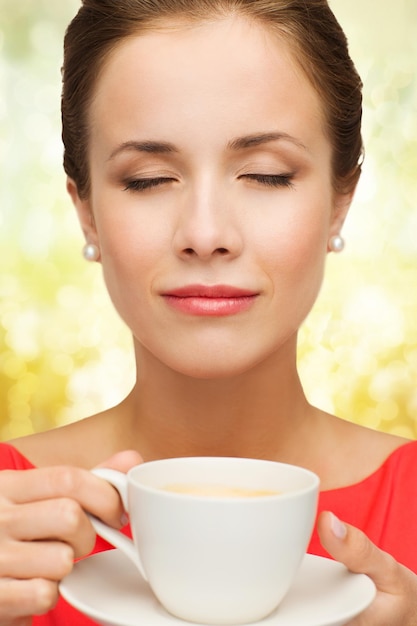 Freizeit-, Glücks- und Getränkekonzept - lächelnde Frau in rotem Kleid mit geschlossenen Augen, die eine Tasse Kaffee über goldenem Lichthintergrund hält